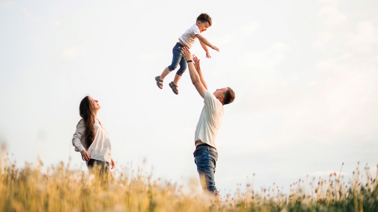 Smiling mother standing beside and happy father lifting up his baby boy in the field; image used for the HSBC International Services Employee Banking Solutions Page.